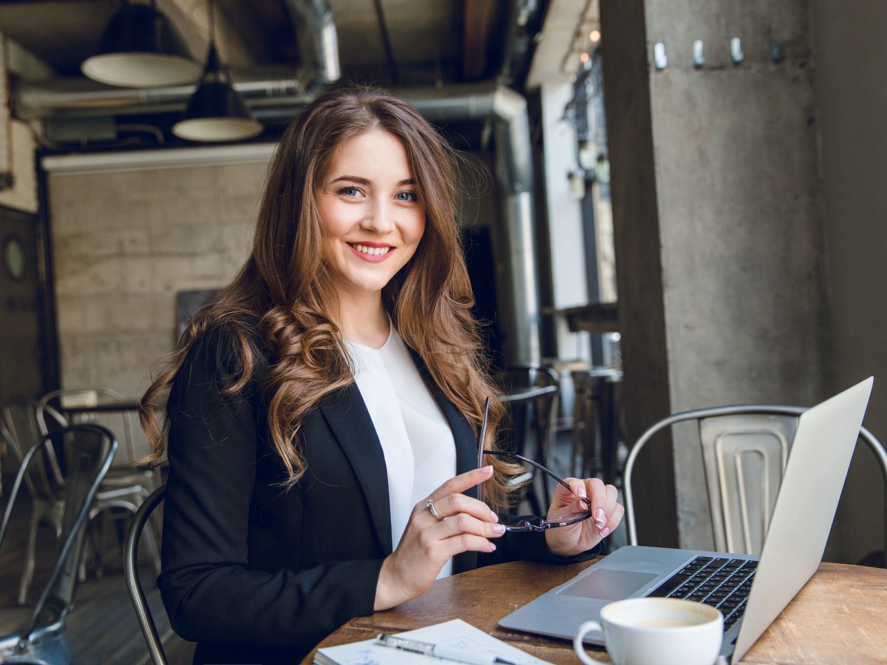 Widely smiling businesswoman working on laptop sitting in a cafe. Woman with long hair wears black jacket and white blouse and holds black eyeglasses in hands.